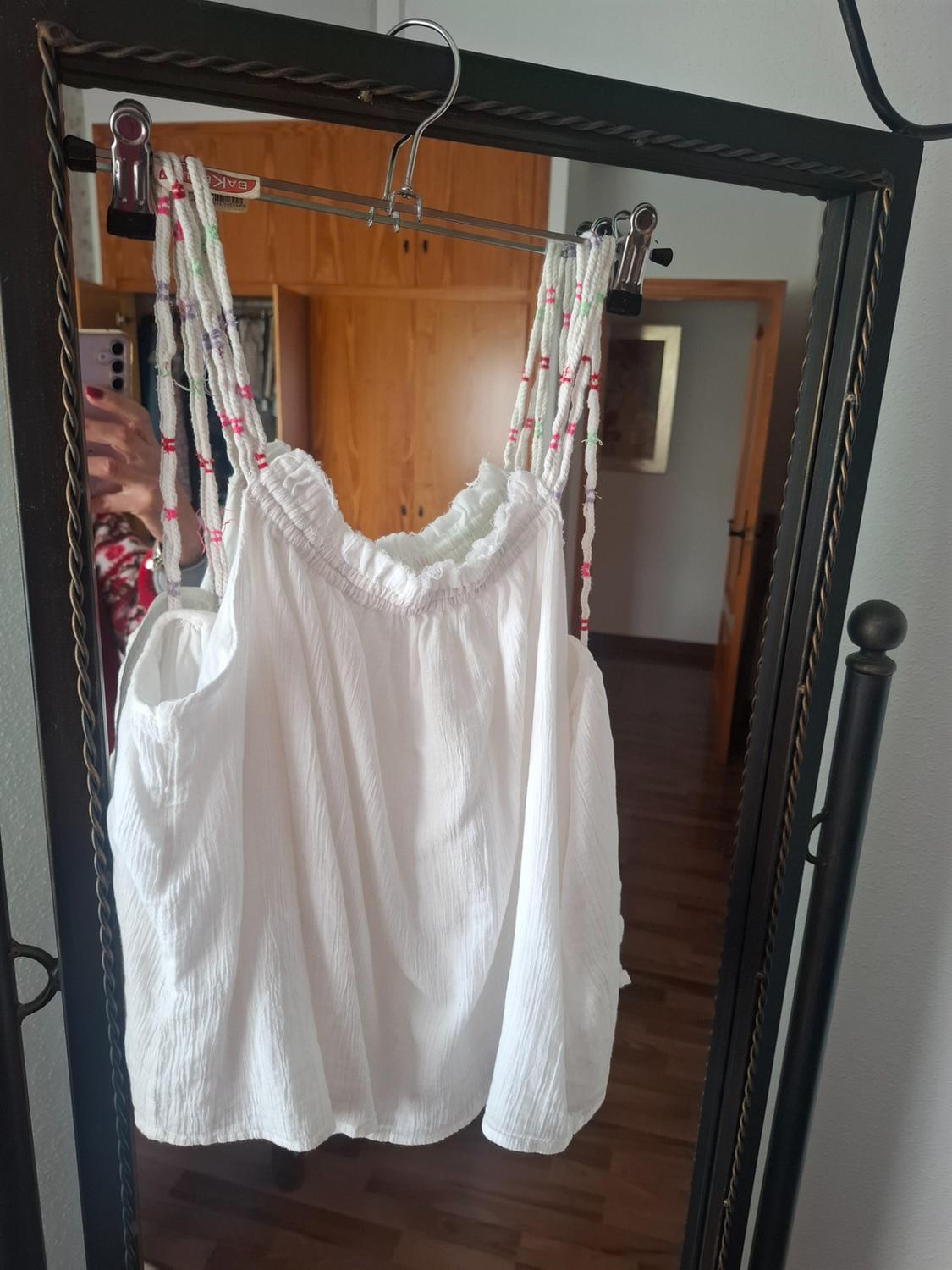 White blouse hanging on a mirror with colourful string straps. Reflection of a person taking the photo in a bedroom.