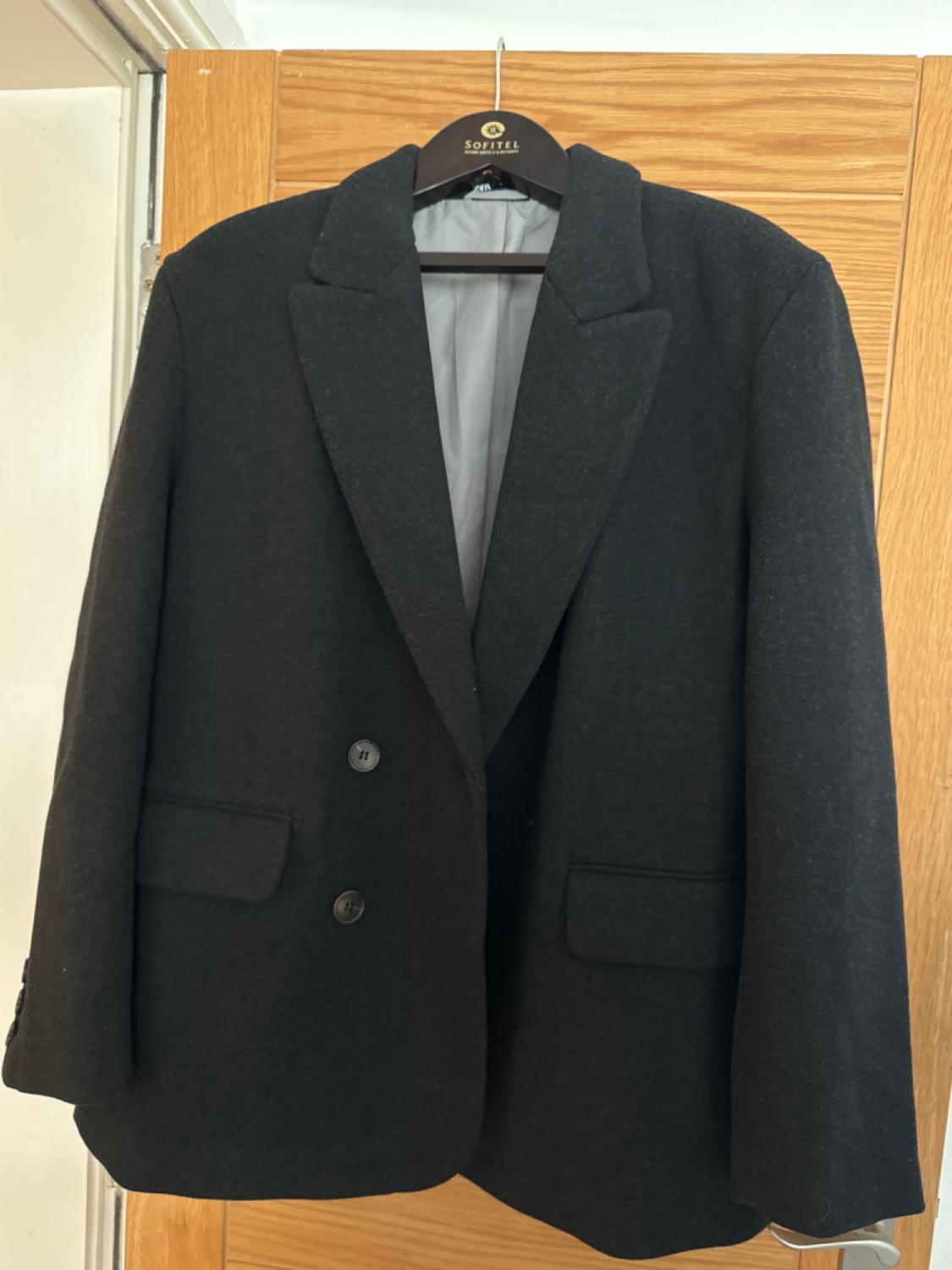 Black jacket hanging on a Sofitel hanger, with two buttons and classic lapels, on a light wooden door.