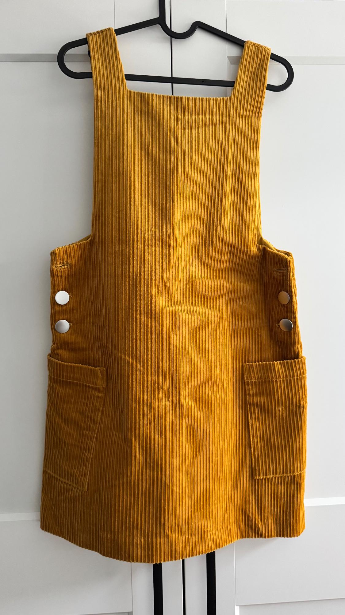Mustard corduroy pinafore dress with wide straps, silver side buttons and front pockets, hanging on a black hanger.