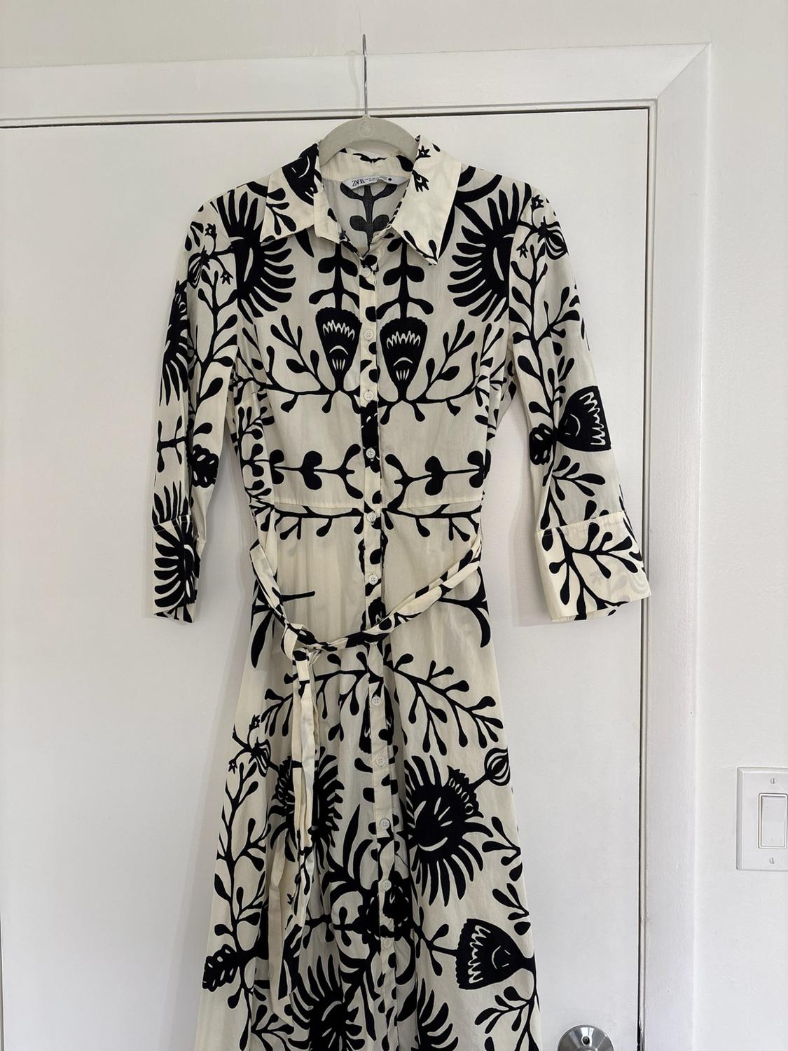 White shirt dress with a black floral print, buttons and belt, hanging on a white door. ZARA label.