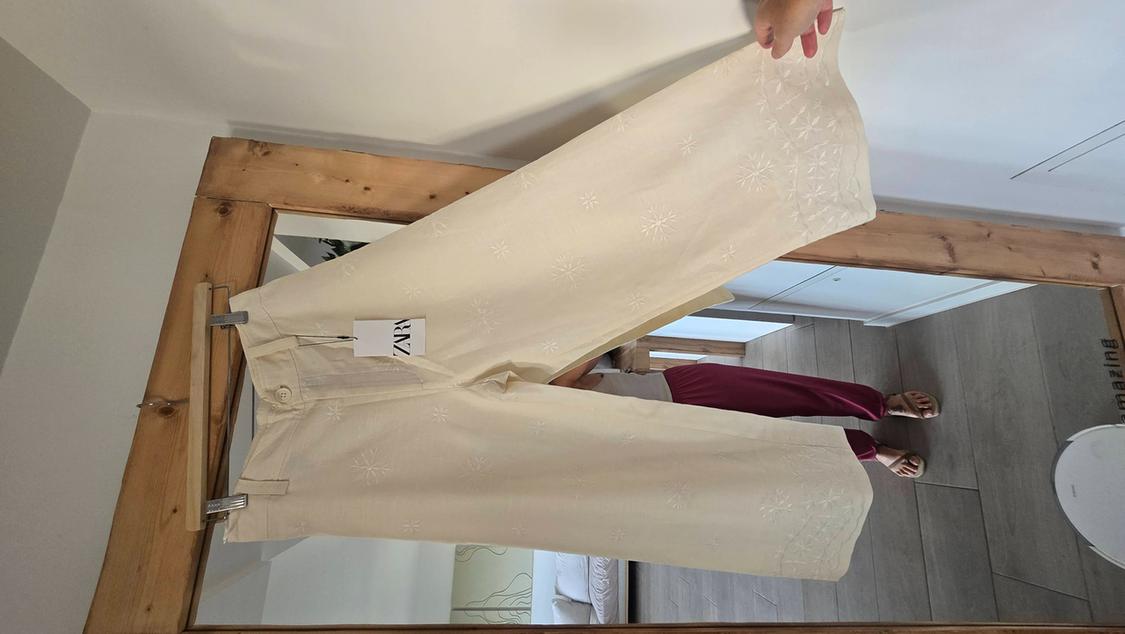 Cream floral trousers hanging on a wooden mirror. Reflection of a person in burgundy trousers and sandals. Visible label: "ZARA".