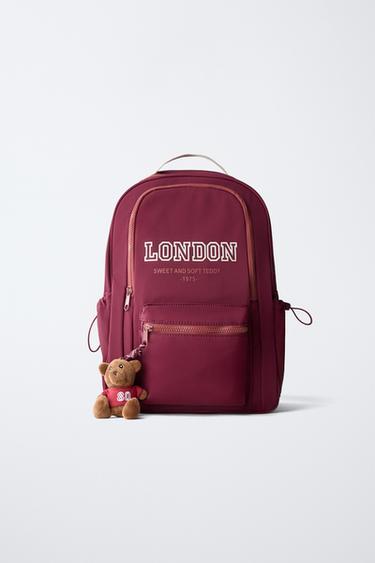 Frontansicht eines bordeauxroten Rucksacks mit dem Text "LONDON SWEET AND SOFT TEDDY 1975", Vordertasche, Bären-Charm und Seitentaschen.
