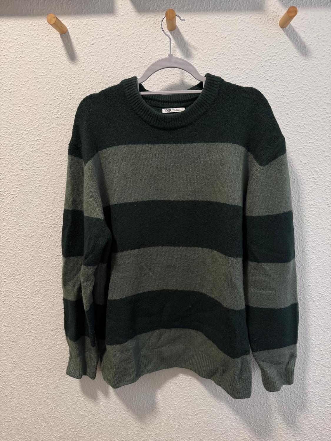 Wide-striped jumper in dark and light green tones, hanging on a white hanger against a white wall with wooden hooks.