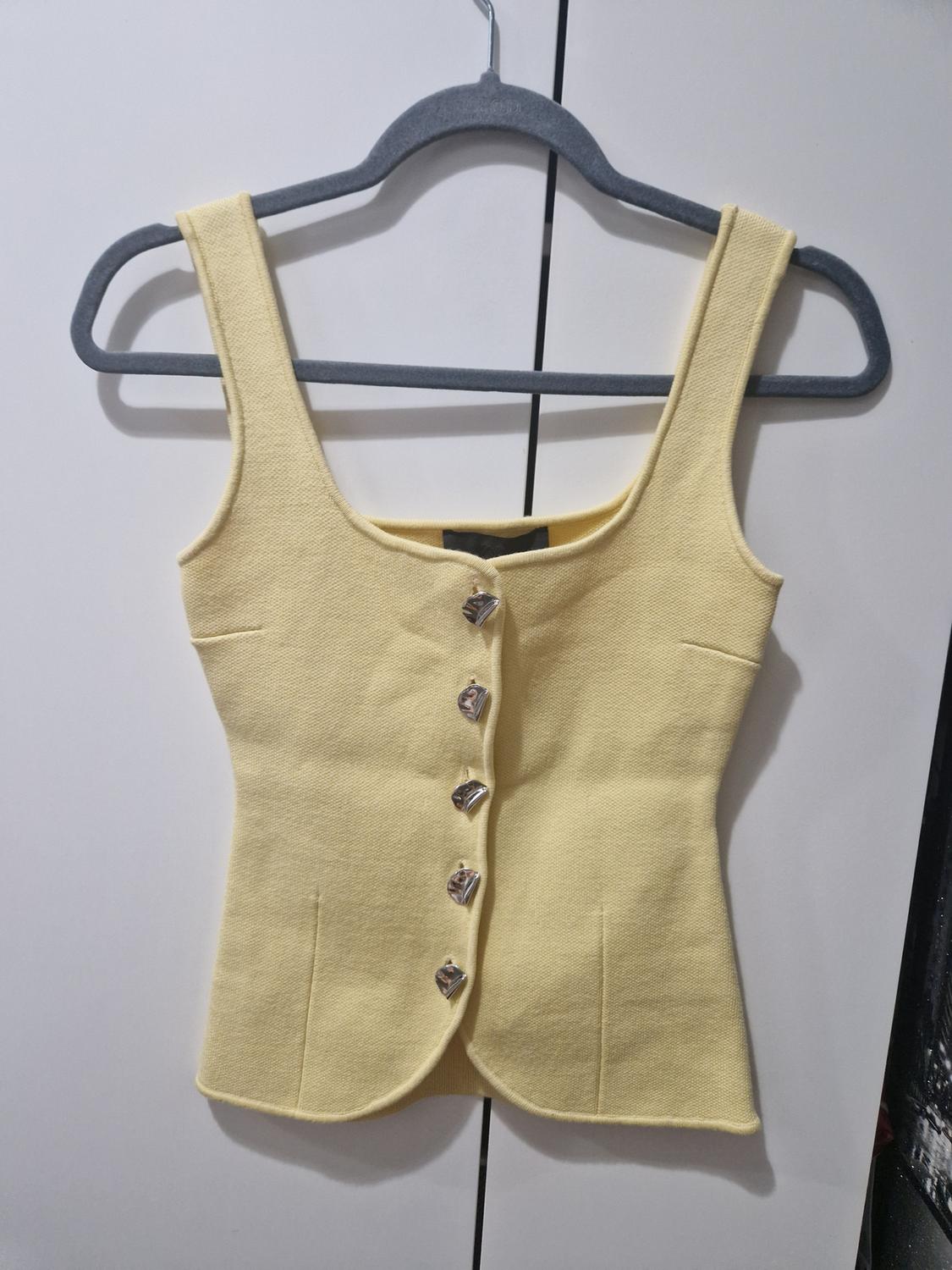Pale yellow sleeveless top with a square neckline and silver-coloured button fastening on the front, hanging on a grey hanger against a white background.