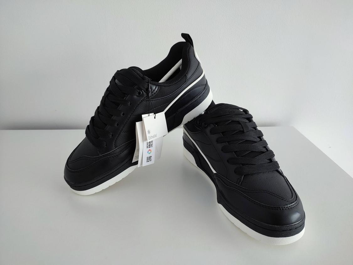 Black trainers with laces and a white sole on a white background. Label with the text: Ortholite, MORE COLOURS ONLINE.