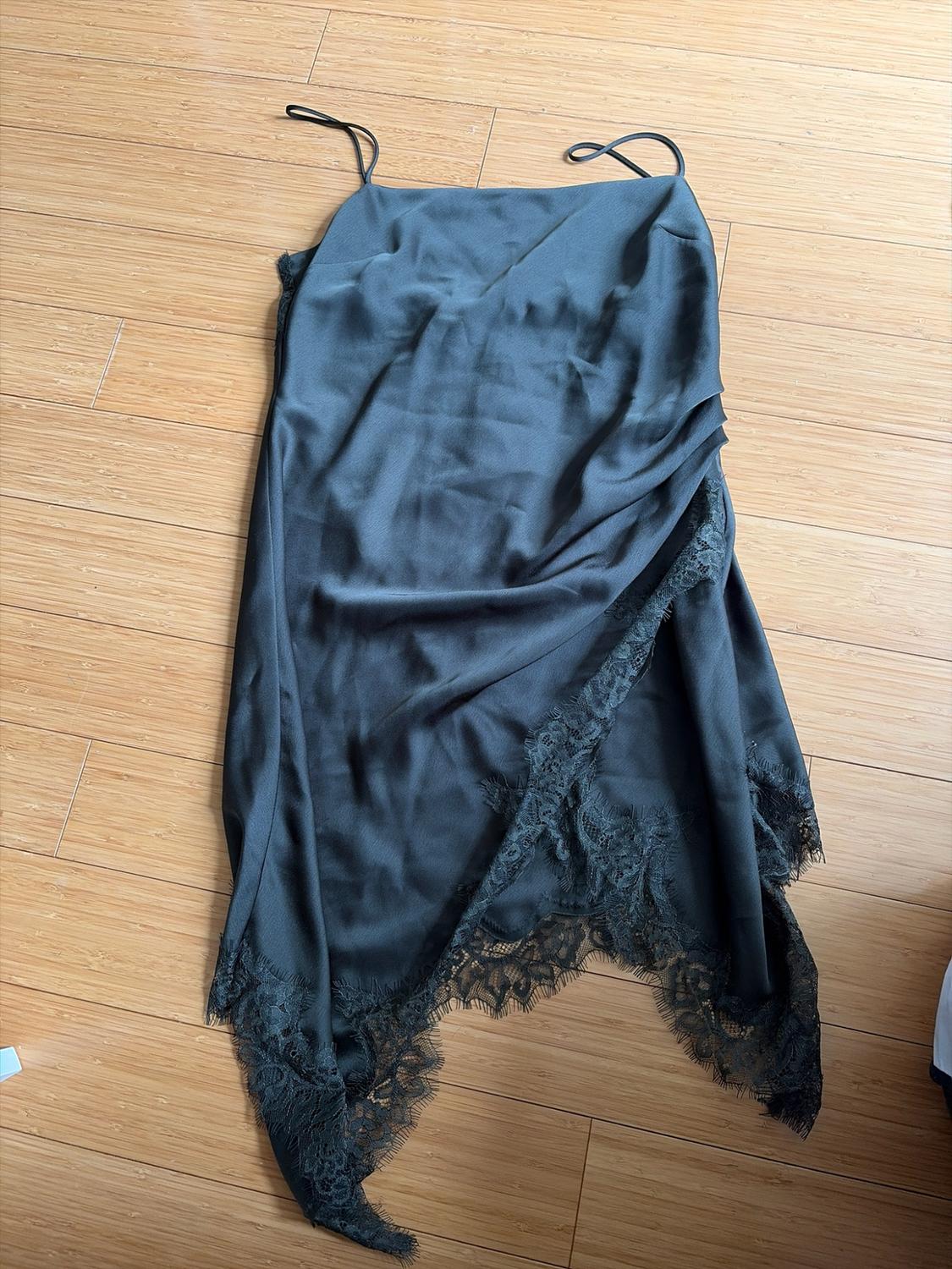 Black satin slip dress with spaghetti straps and an asymmetrical lace hem, laid out on a wooden floor.