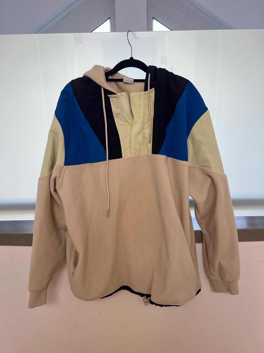 Hoodie hanging on a hanger. Colour-block design in beige, black and blue with mixed textures and adjustable drawstrings.