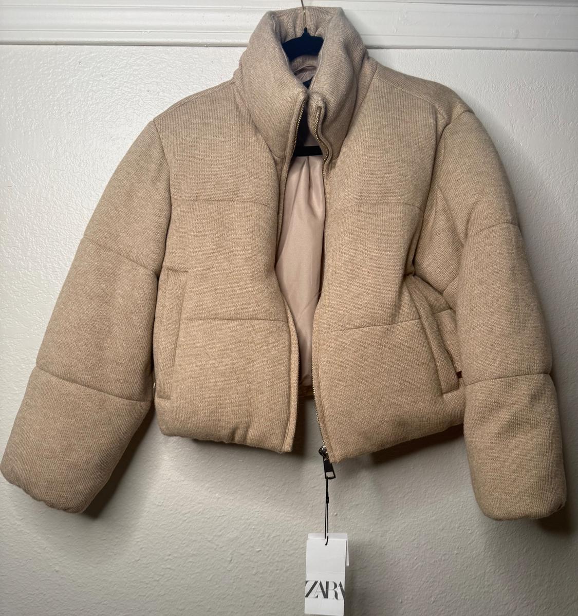 Beige quilted jacket hanging on a black hanger. Visible tag with the text "ZARA". White wall background.