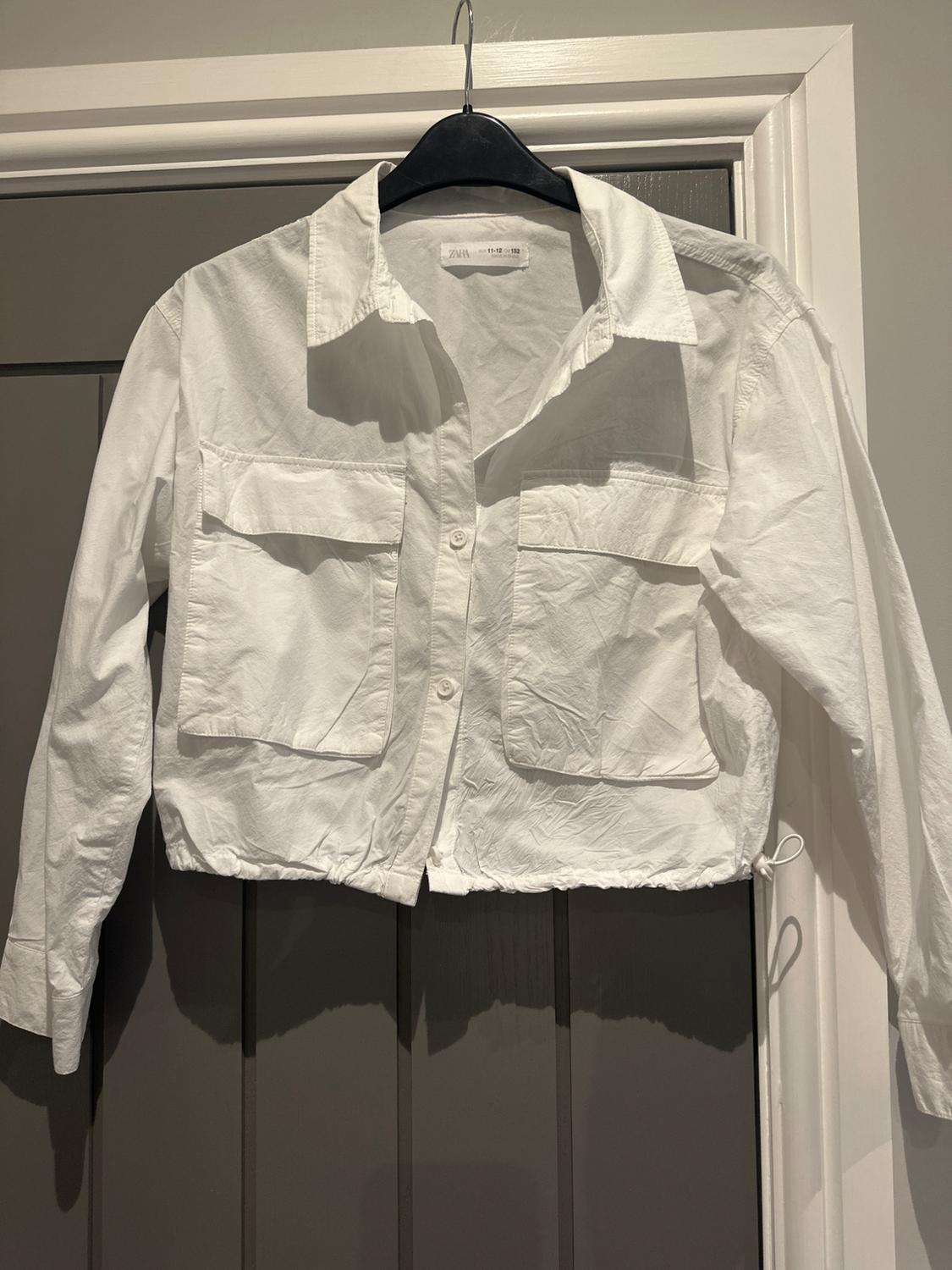 Long-sleeved white shirt with front pockets, hanging on a black coat hanger over a grey door. Label visible on the collar.
