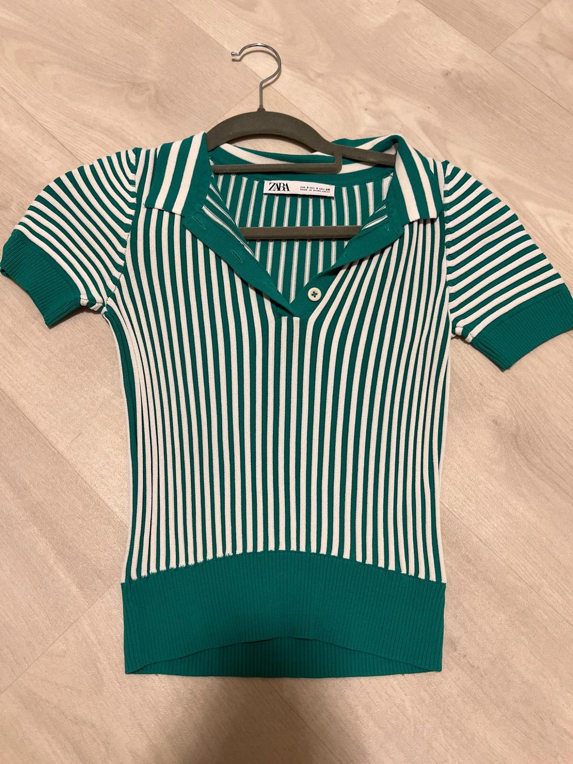 Short-sleeved polo shirt with green and white stripes, buttoned collar and green hem. Hanging on a coat hanger above a wooden floor. Visible text: ZARA.