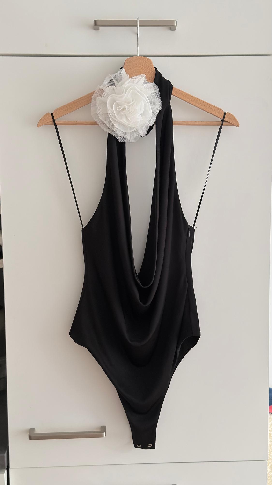 Black bodysuit with thin straps and a white flower detail on the shoulder, hanging on a wooden hanger against a white background.