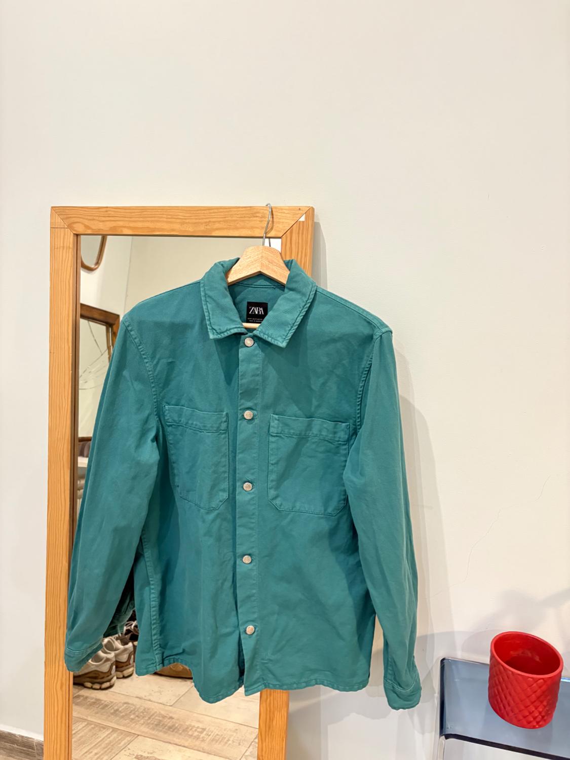 Green shirt hanging on a wooden mirror, with a red vase to the right and shoes reflected in the mirror.
