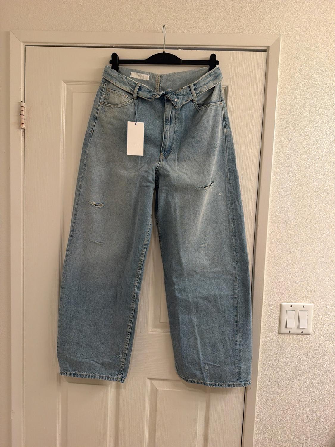 Light blue wide-leg jeans with rips and a crossover waist, hanging on a hanger over a white door. Casual and relaxed vibe.