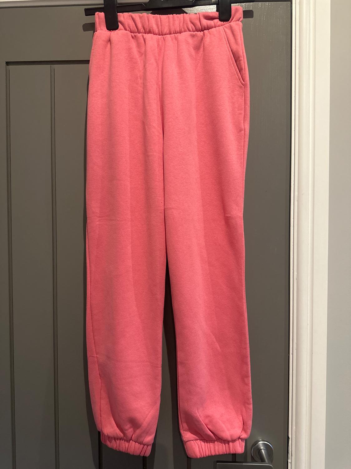 Pink jogging bottoms hanging on a grey door. They have an elasticated waistband and fitted cuffs.