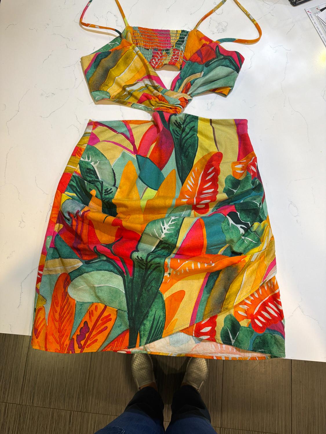 Top and skirt set with a colorful tropical print on a white background. Person with boots visible at the bottom.