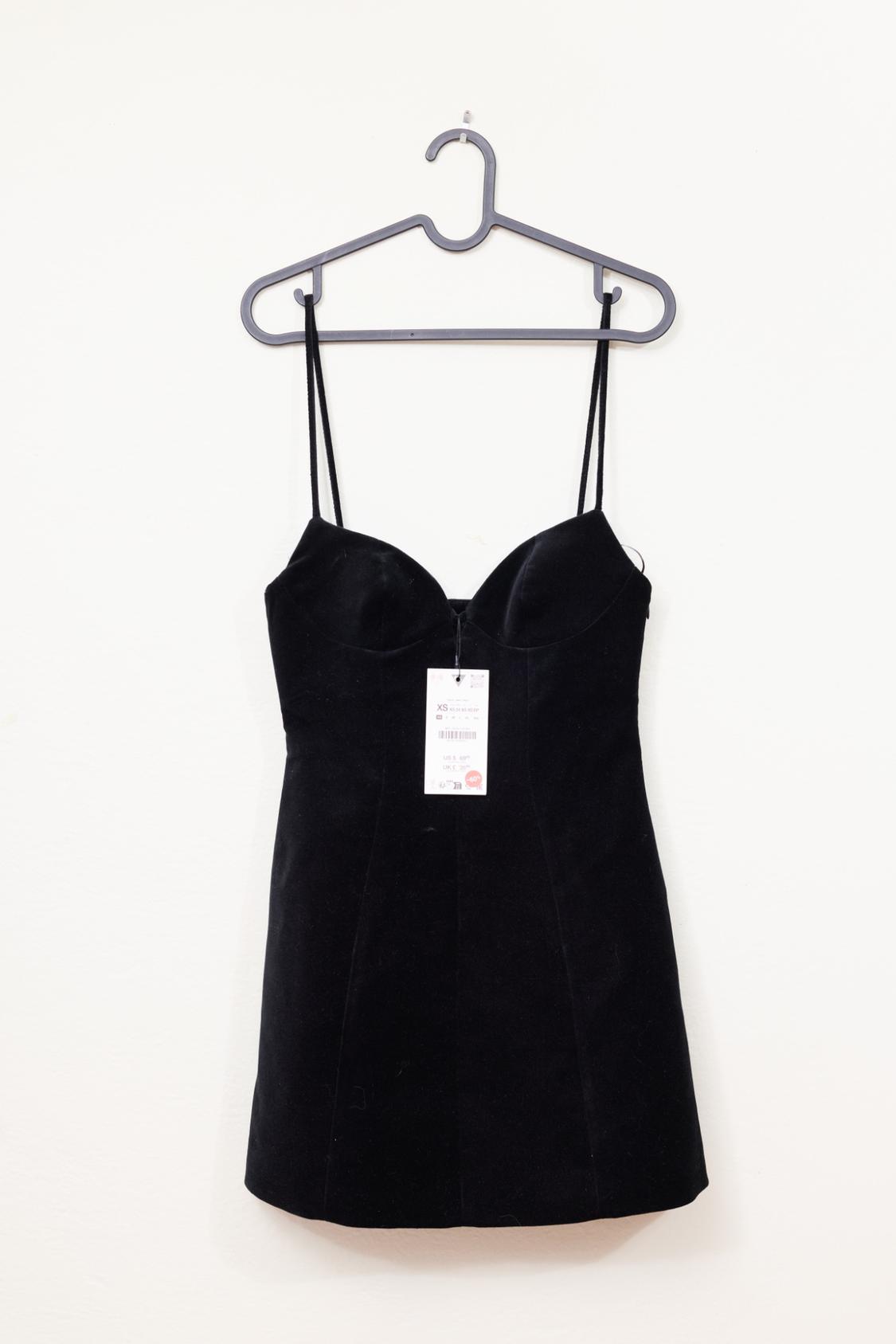 Short black dress with thin straps, lingerie-style, hanging on a gray hanger against a white background. It has a price tag in the center.