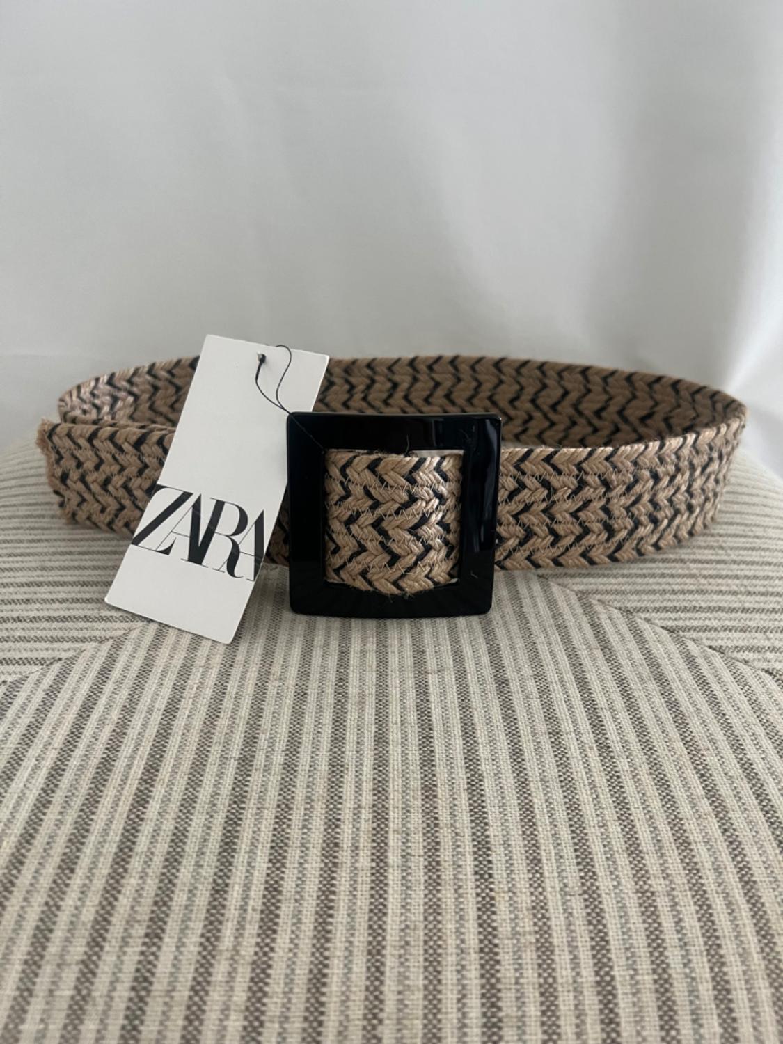 Beige braided belt with black zigzag and black square buckle. Label with the text ZARA. Rests on striped fabric.