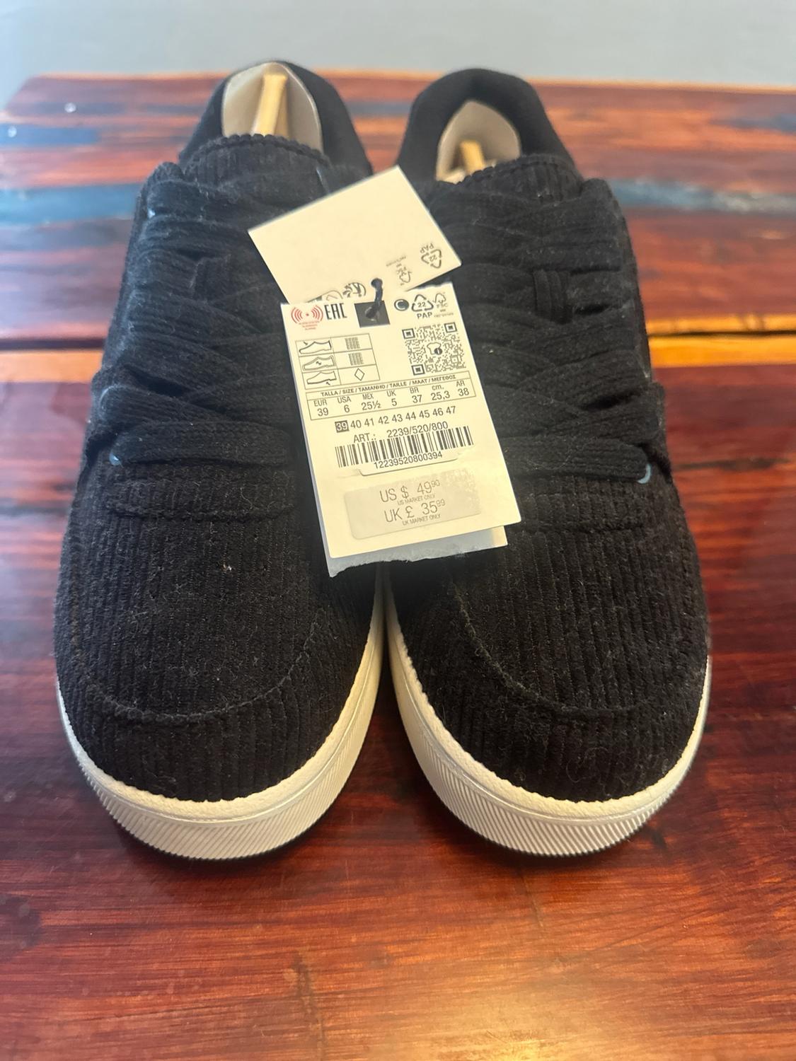 Pair of black corduroy trainers with a white sole on a wooden surface. Label with text: US $ 49.90 UK £ 35.99.