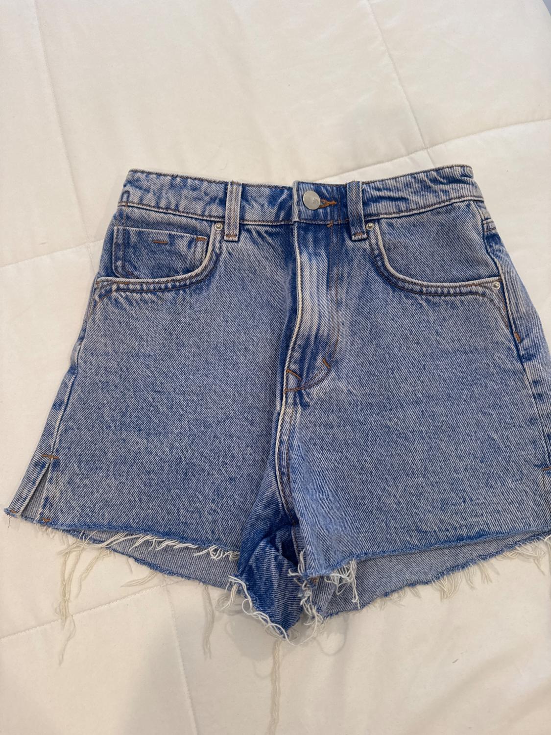 Blue denim shorts with frayed hem, front pockets and belt loops, on a white surface.