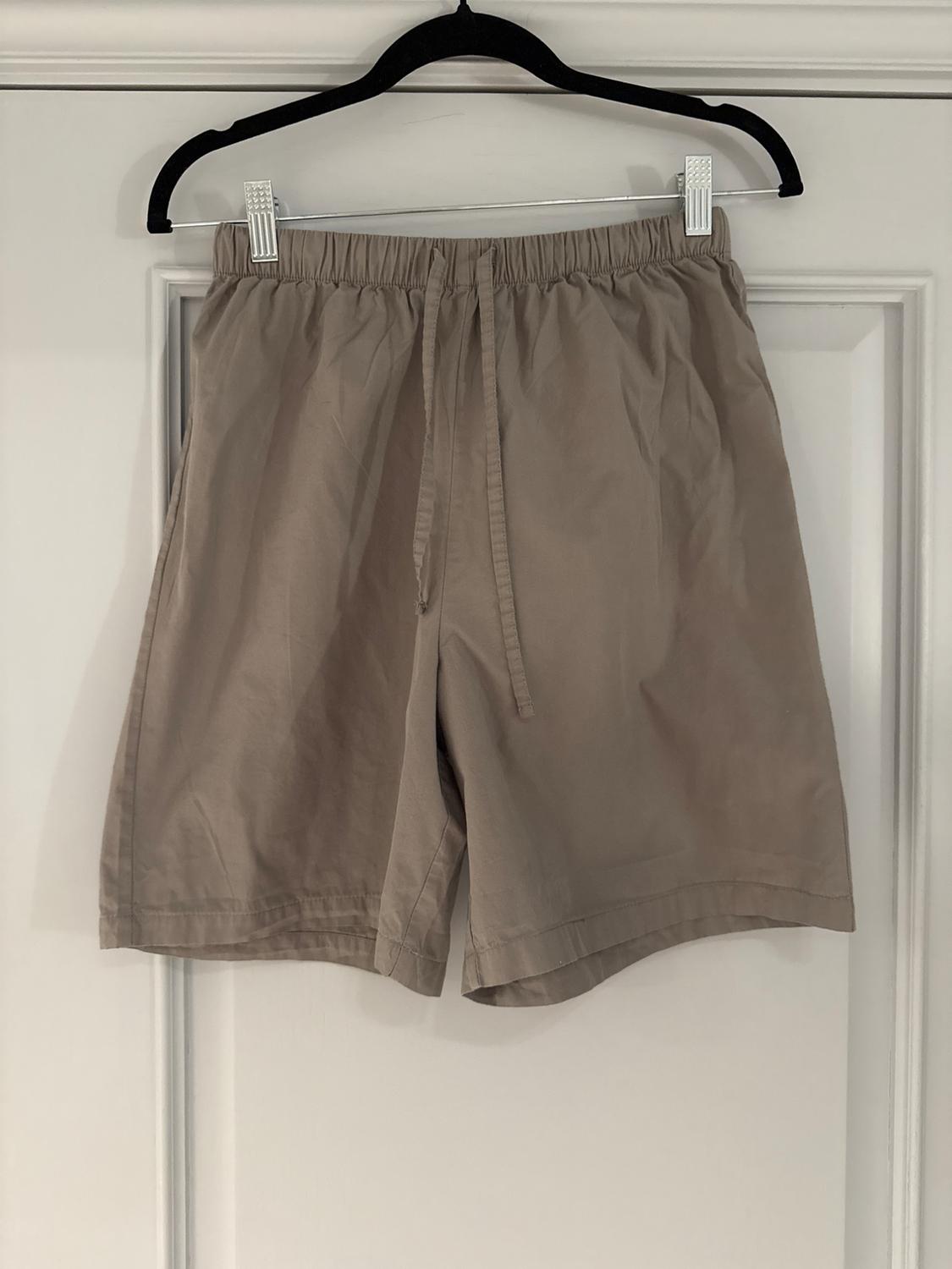 Beige shorts hung on a black hanger over a white door. Elasticated waist with adjustable drawstring.
