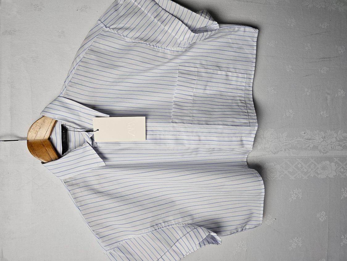 White shirt with blue stripes hanging on a wooden hanger against a light surface. White label visible on the collar.
