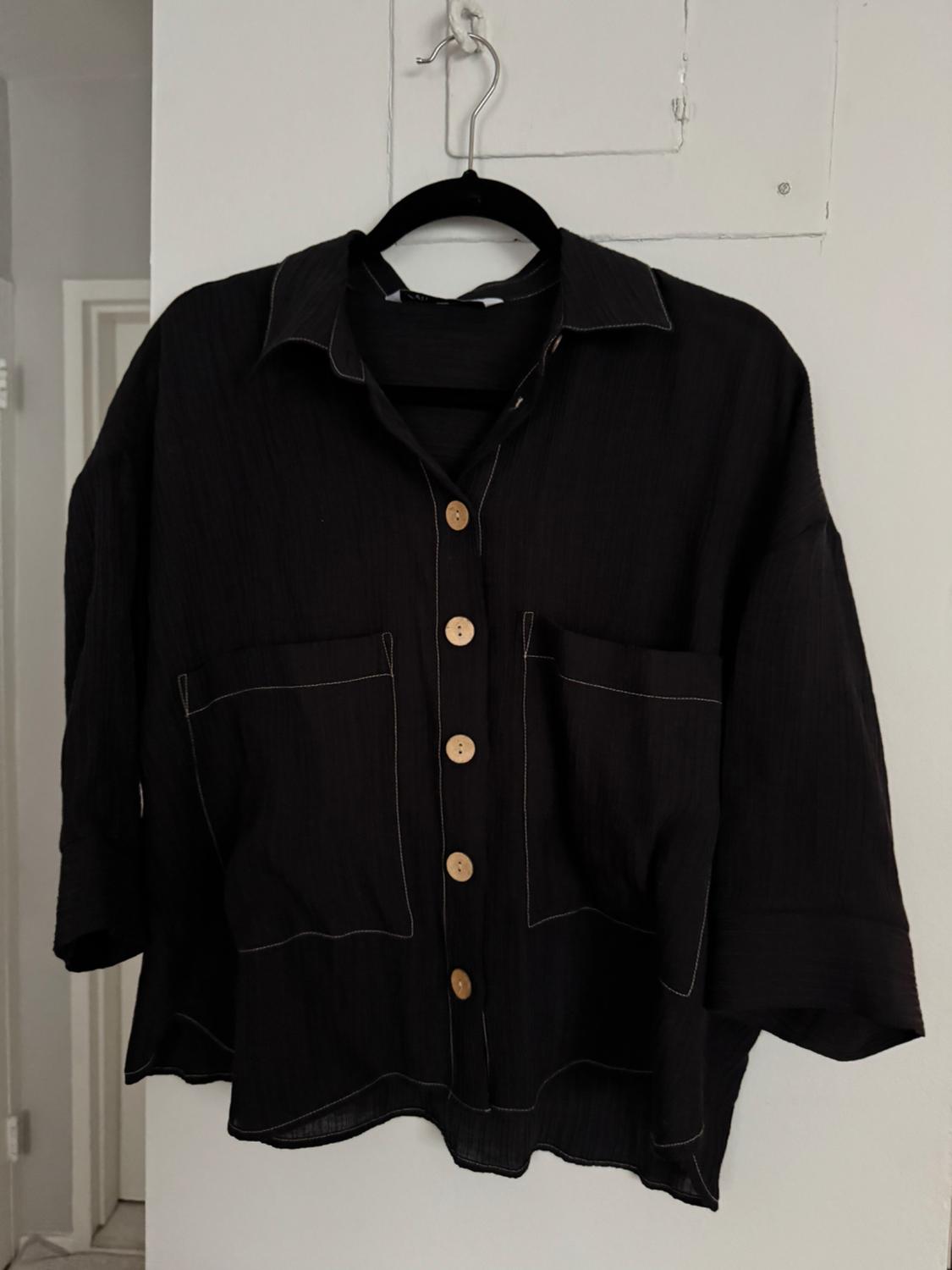 Long-sleeved black shirt with brown buttons and visible stitching, hanging on a black hanger against a white wall.