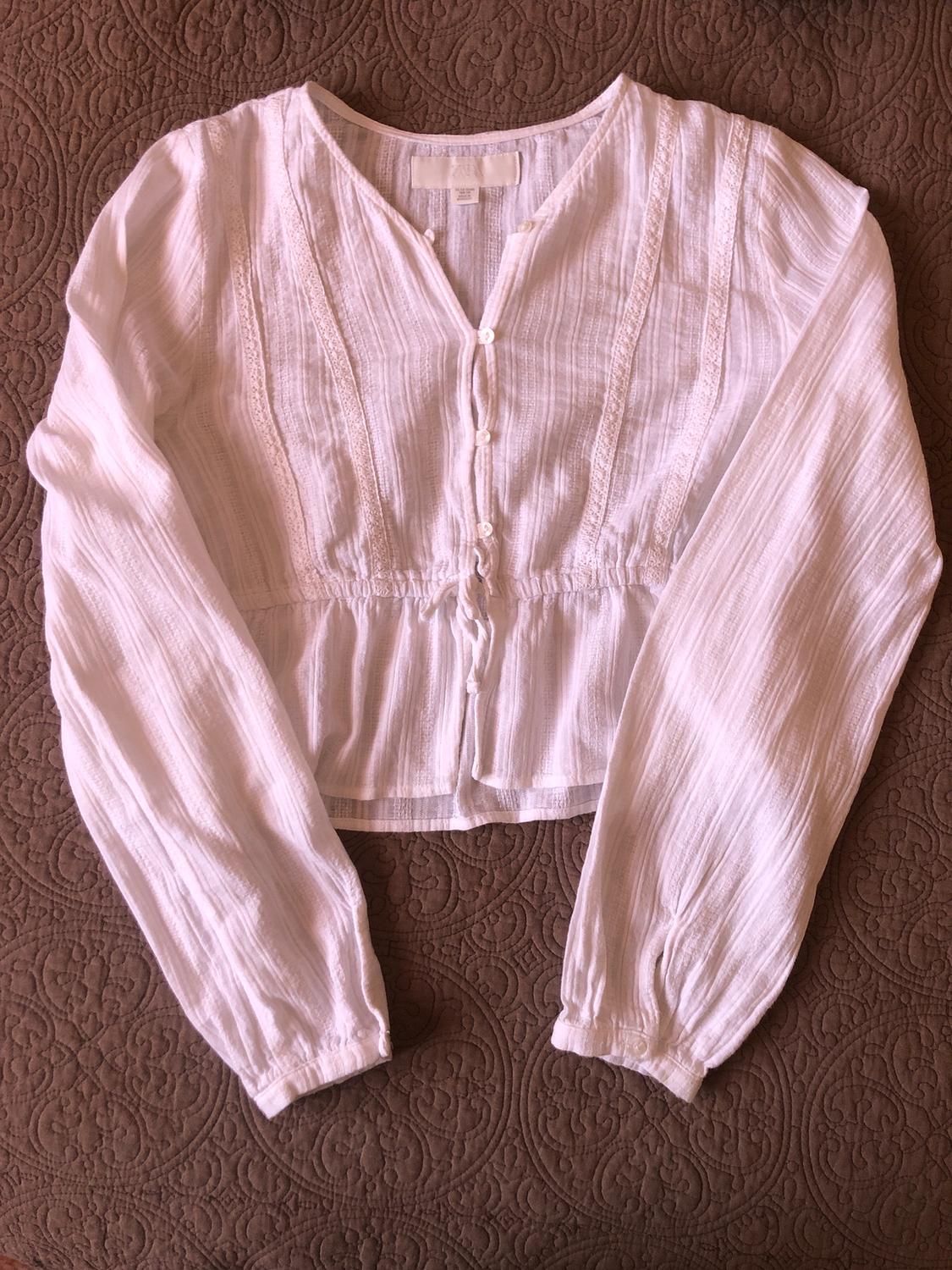 Long-sleeved white blouse with front buttons and lace details, on a brown bedspread with a geometric pattern design.