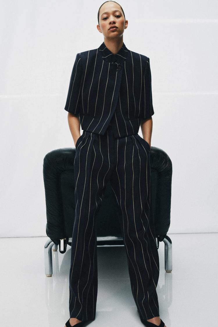 Souprava tmav� modr�ho saka a kalhot s vertik�ln�mi pruhy. Kr�tk� sako se zap�n�n�m na knofl�k, kalhoty se st�edn� vysok�m pasem a bo�n�mi kapsami.