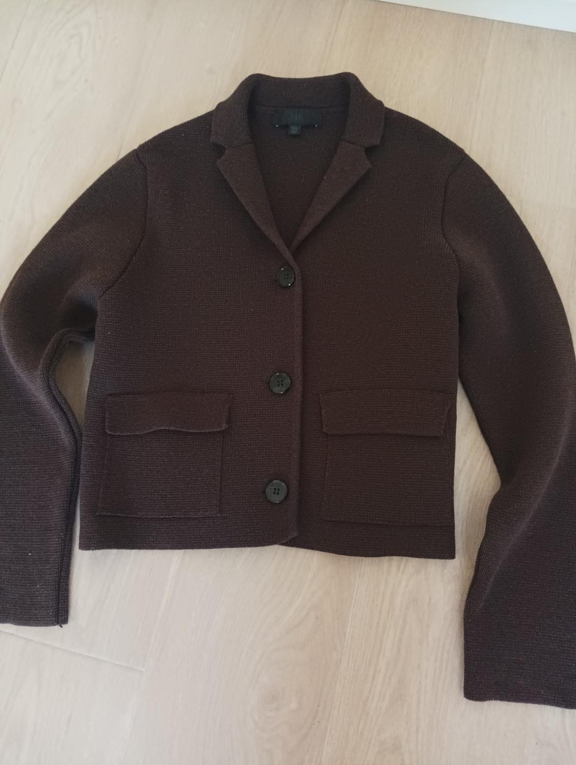 Brown knitted jacket with lapels and black buttons, two front pockets, on a light wooden floor.