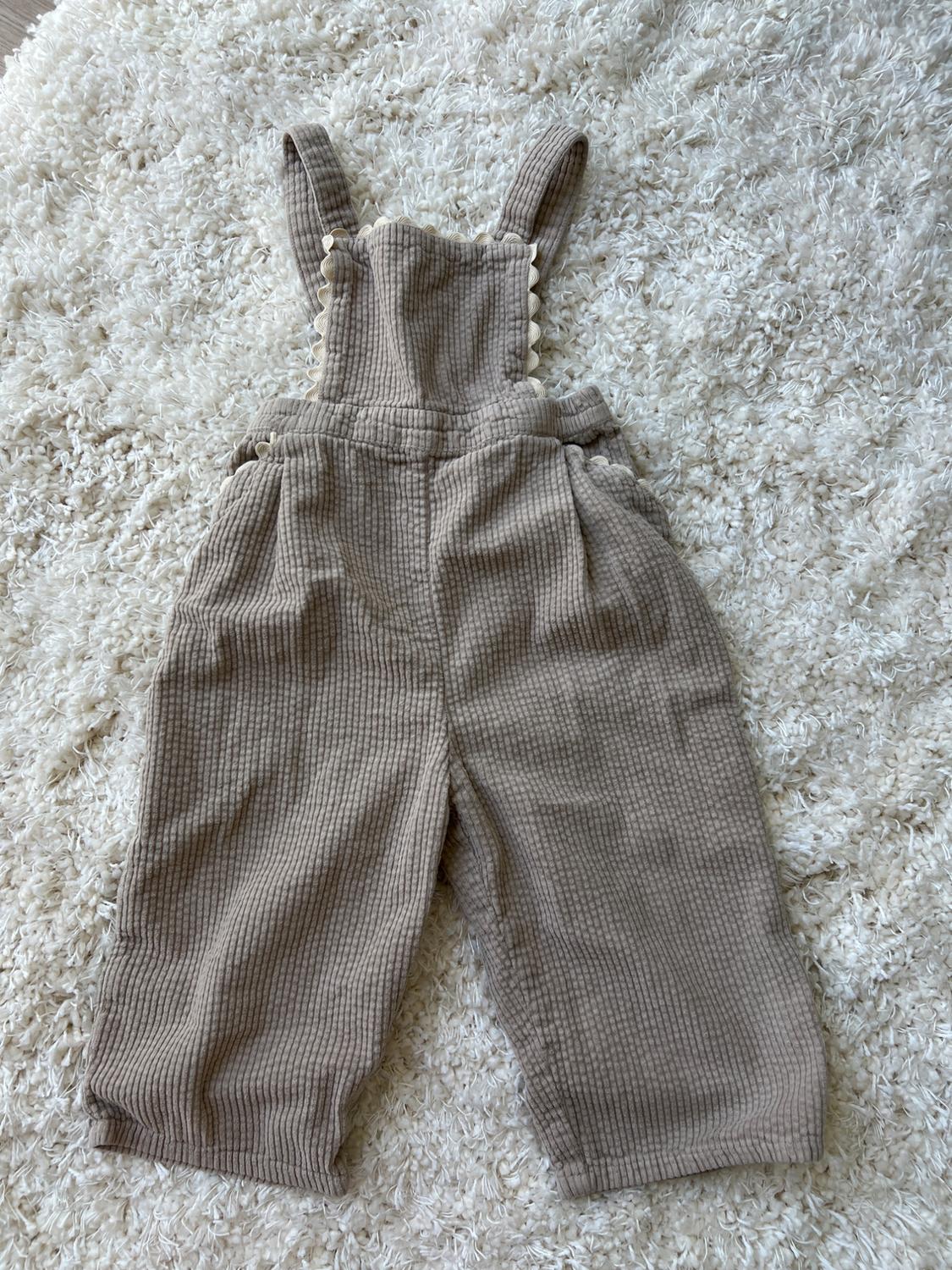Beige ribbed children's dungarees with a wavy trim, on a white shag rug. Relaxed and cosy composition.