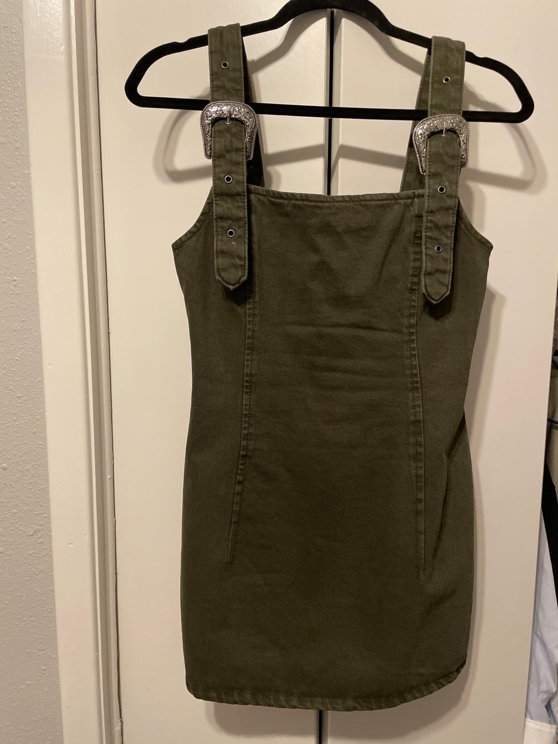 Olive green dress with wide straps and silver buckles, hanging on a black hanger in front of a white door.