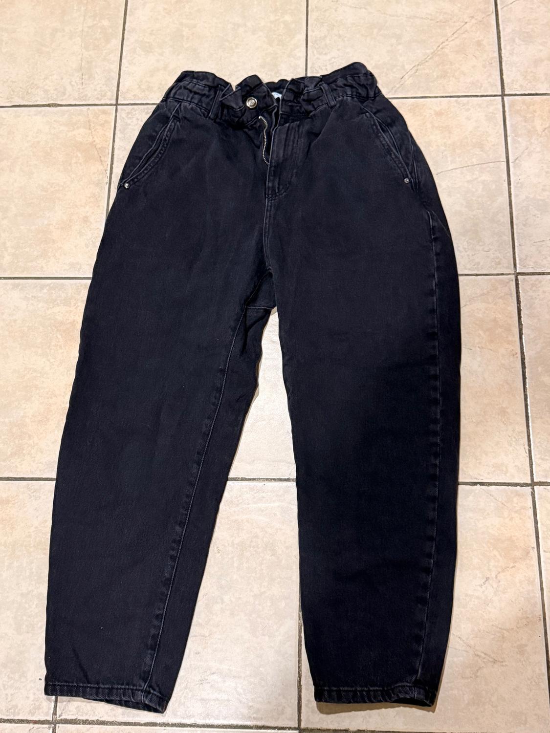 Black denim pants with an elastic waistband and button, placed on a beige tiled floor.
