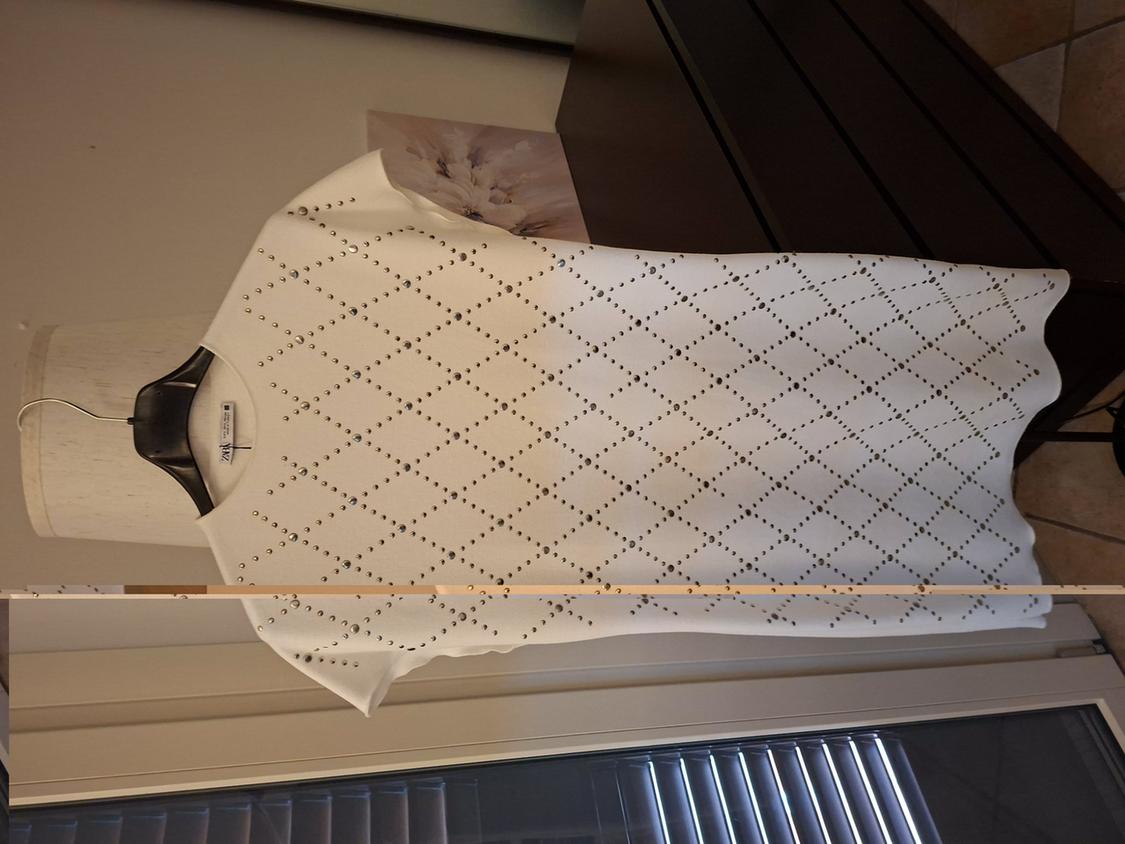 White dress with perforated diamond pattern, hanging on a black hanger against a background of dark furniture and a window.