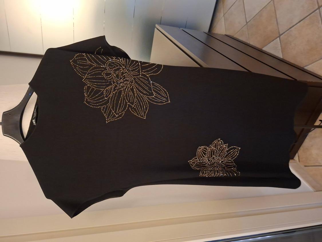 Black dress with gold floral embroidery on the chest and bottom. Hung on a hanger against a background of light-coloured furniture.