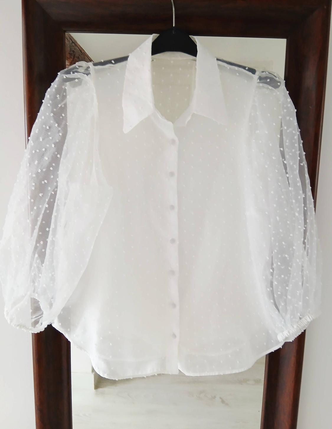 Blouse, white, sheer fabric with polka dots and puff sleeves, hanging on a black hanger against a wooden mirror background.