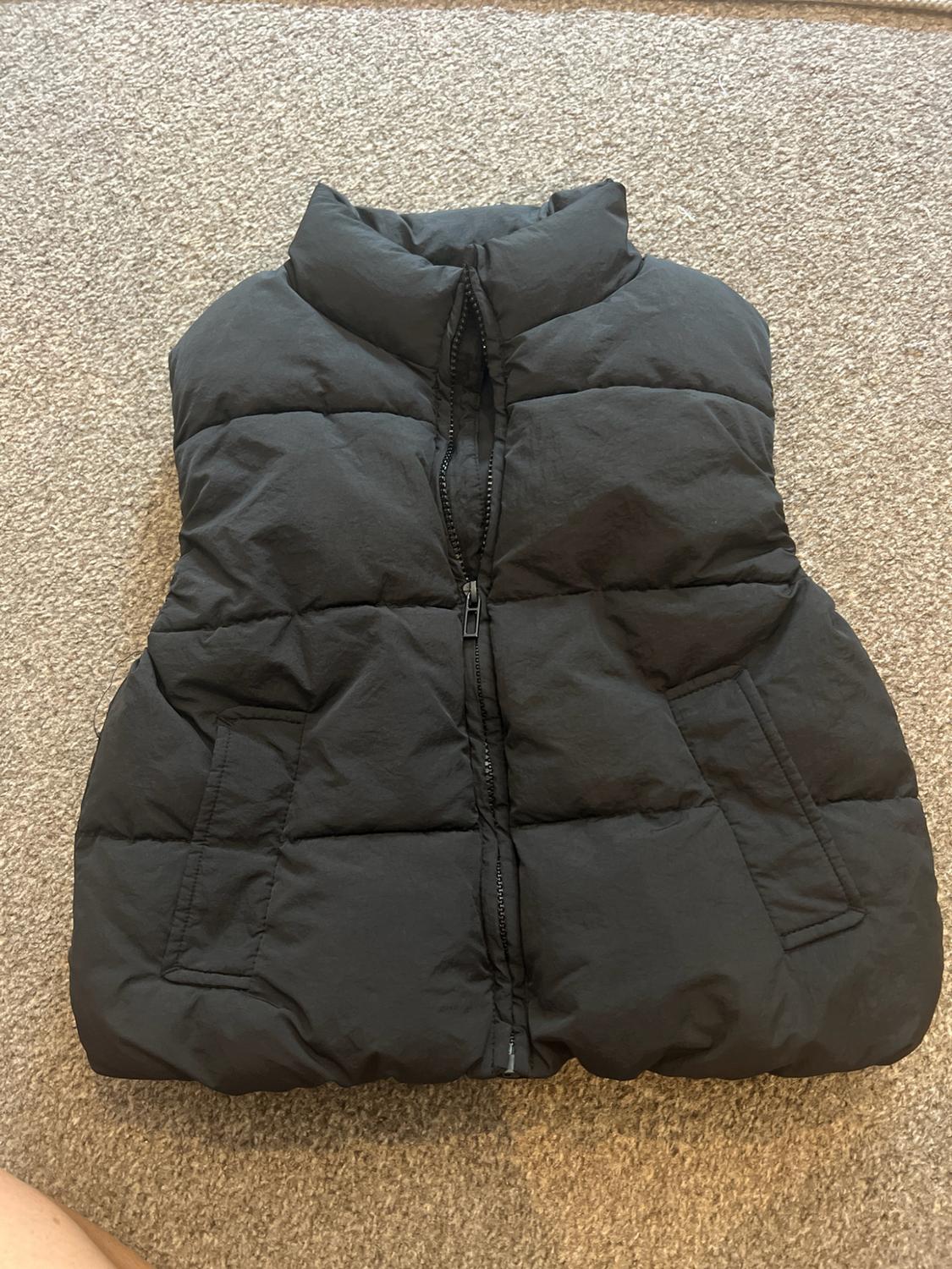 Black high-neck padded gilet with a front zip and side pockets, laid out on a beige carpet.
