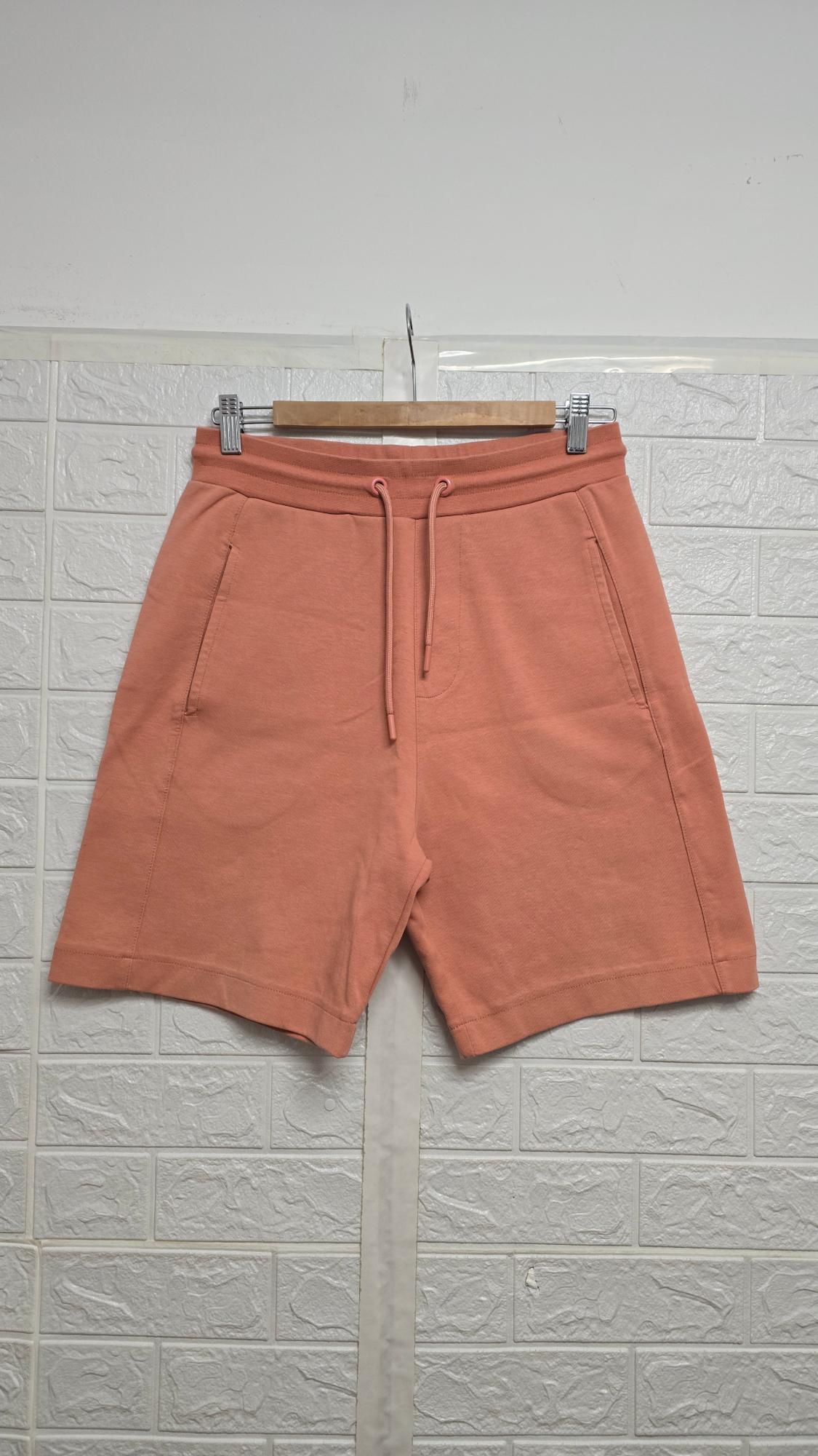 Salmon-coloured shorts hanging on a wooden hanger, with adjustable drawstrings and side pockets, against a white wall background.