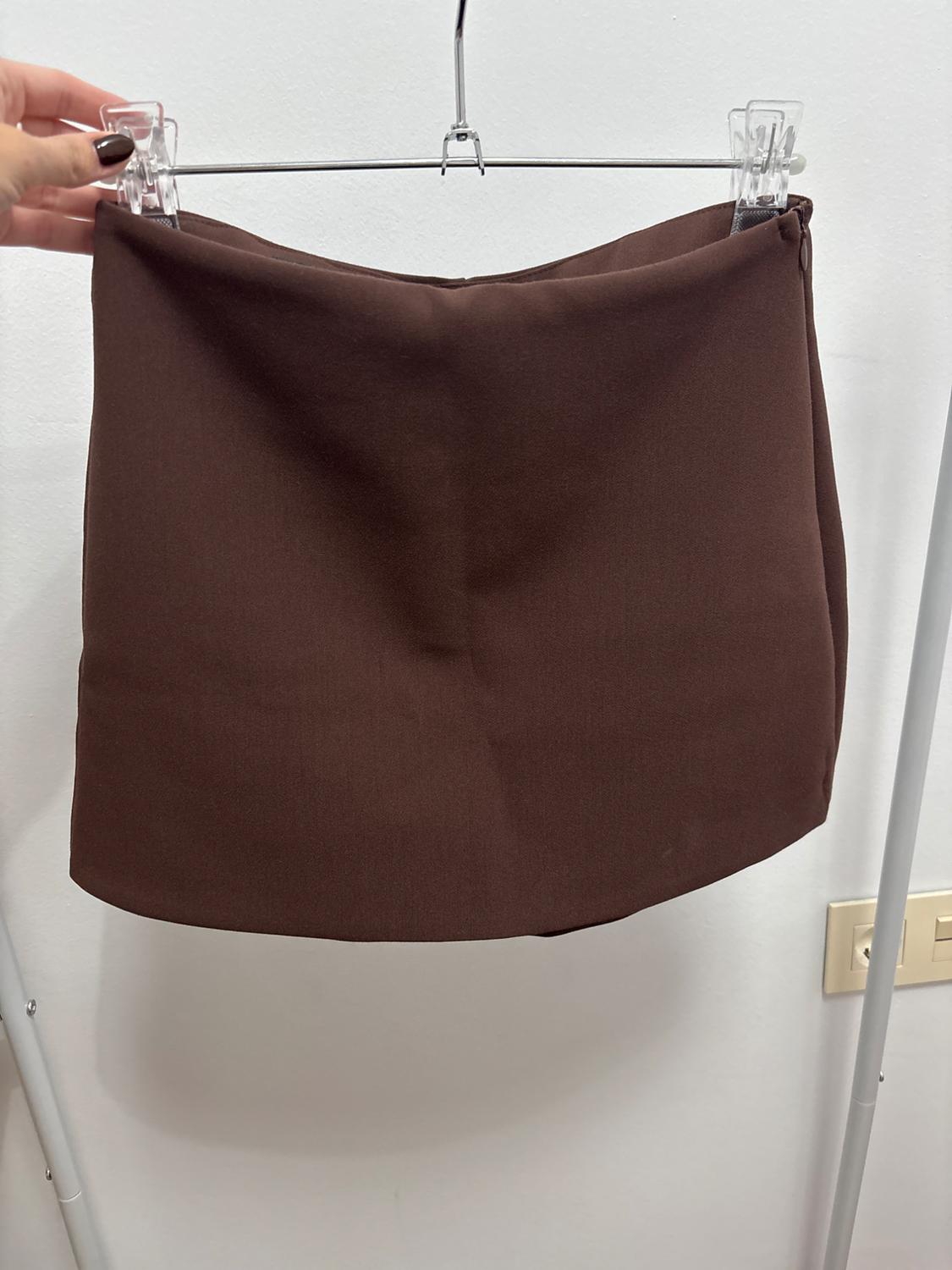A brown skirt is hanging on a metal hanger, held by a hand with dark-painted nails, against a white background.