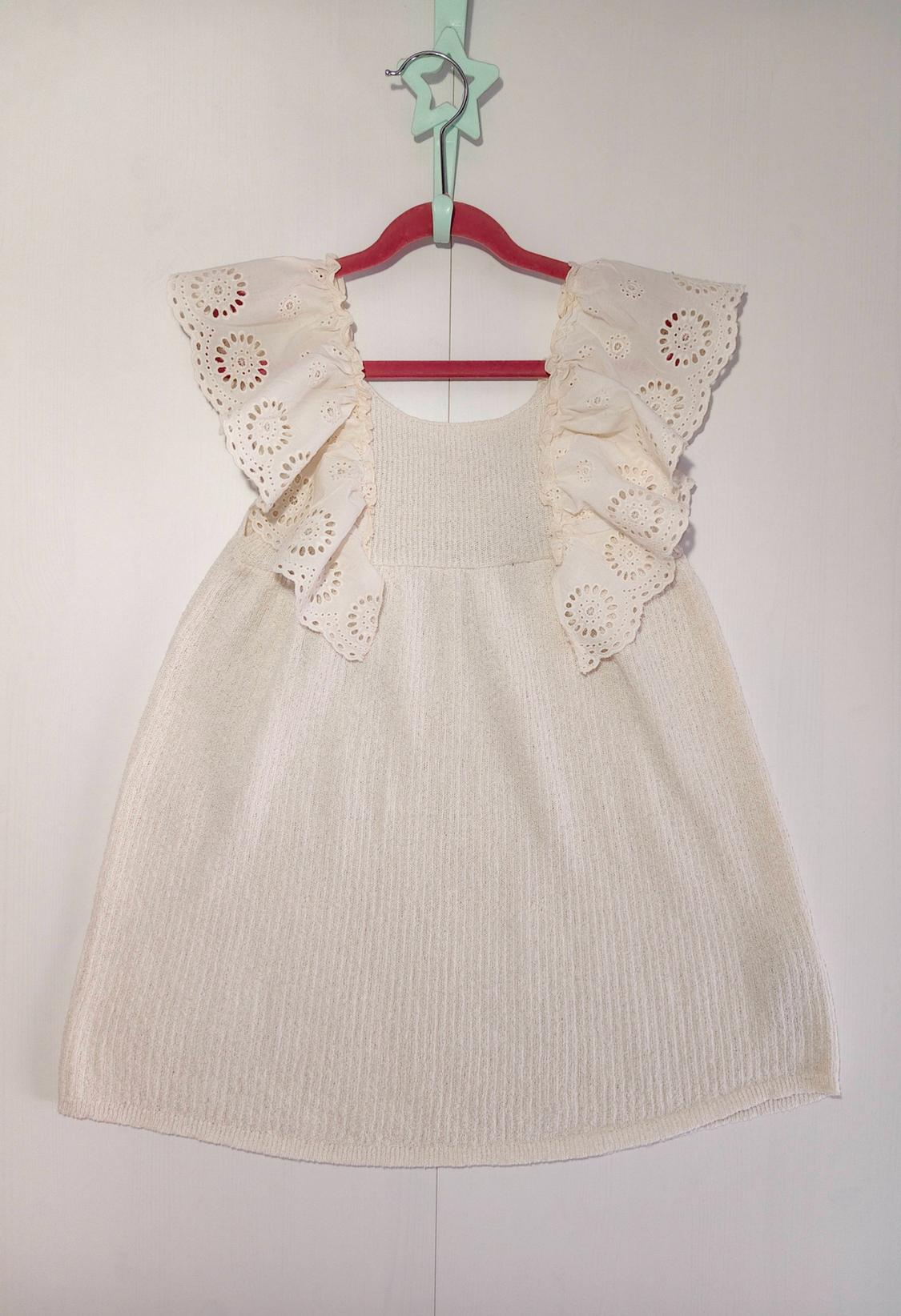 White knitted children's dress with large embroidered ruffles on the sleeves, hanging on a pink hanger against a plain white background.