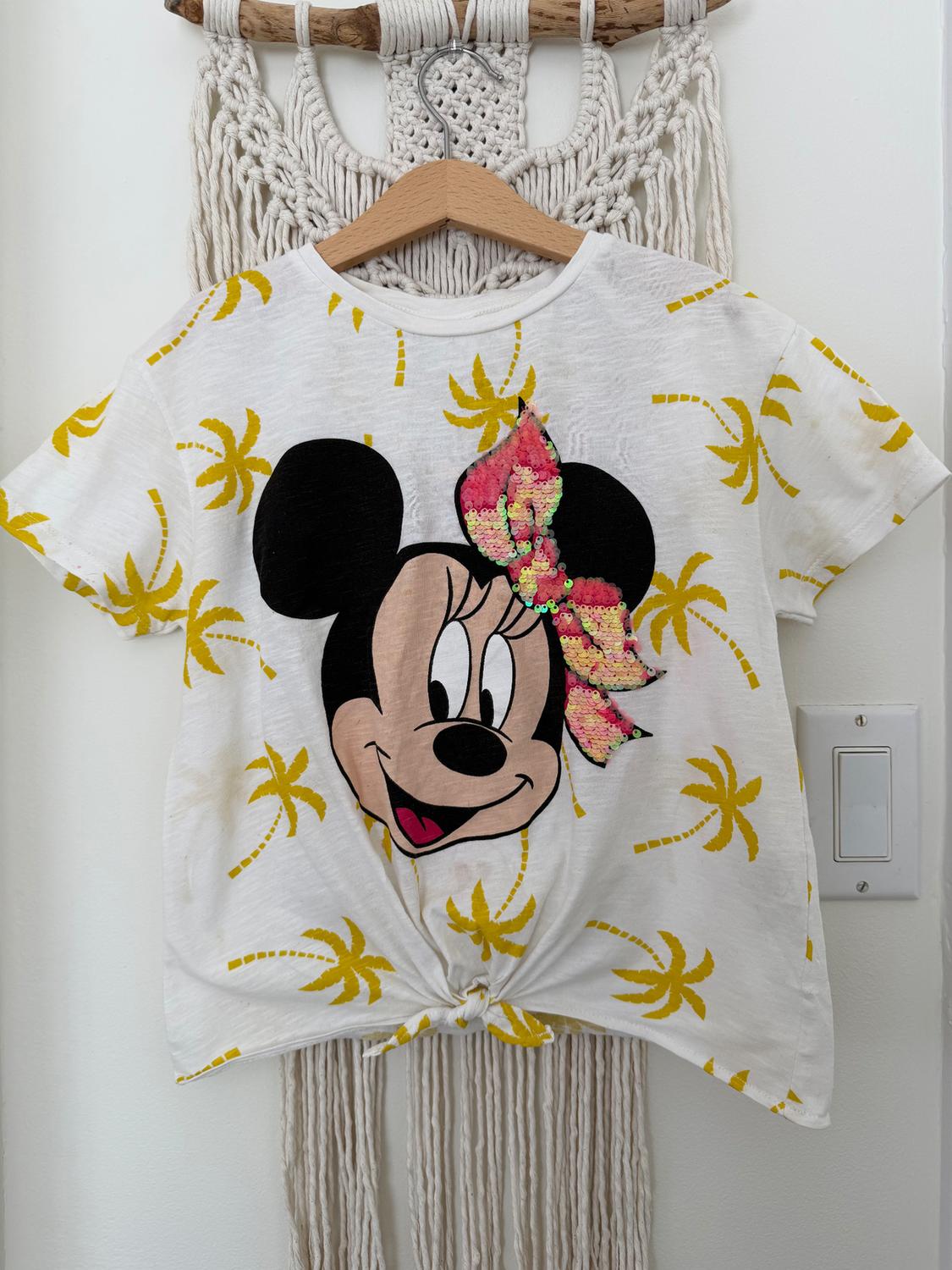 White t-shirt with yellow palm tree print and animated character drawing with sequin bow, hanging on a wooden hanger.