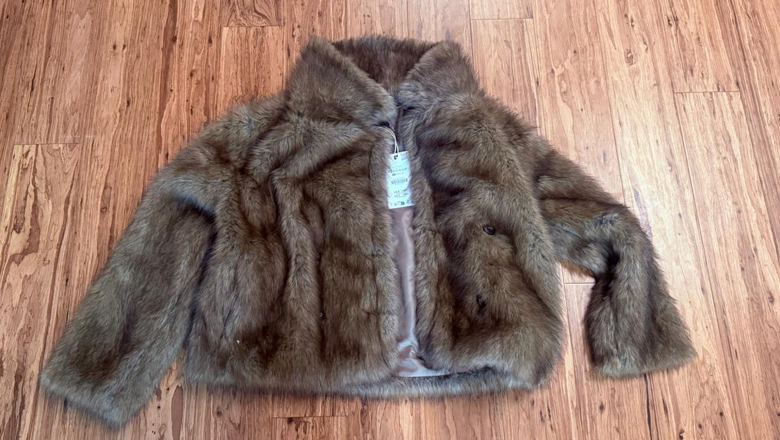 Brown faux fur jacket with tag, open and laid flat on a wooden floor.