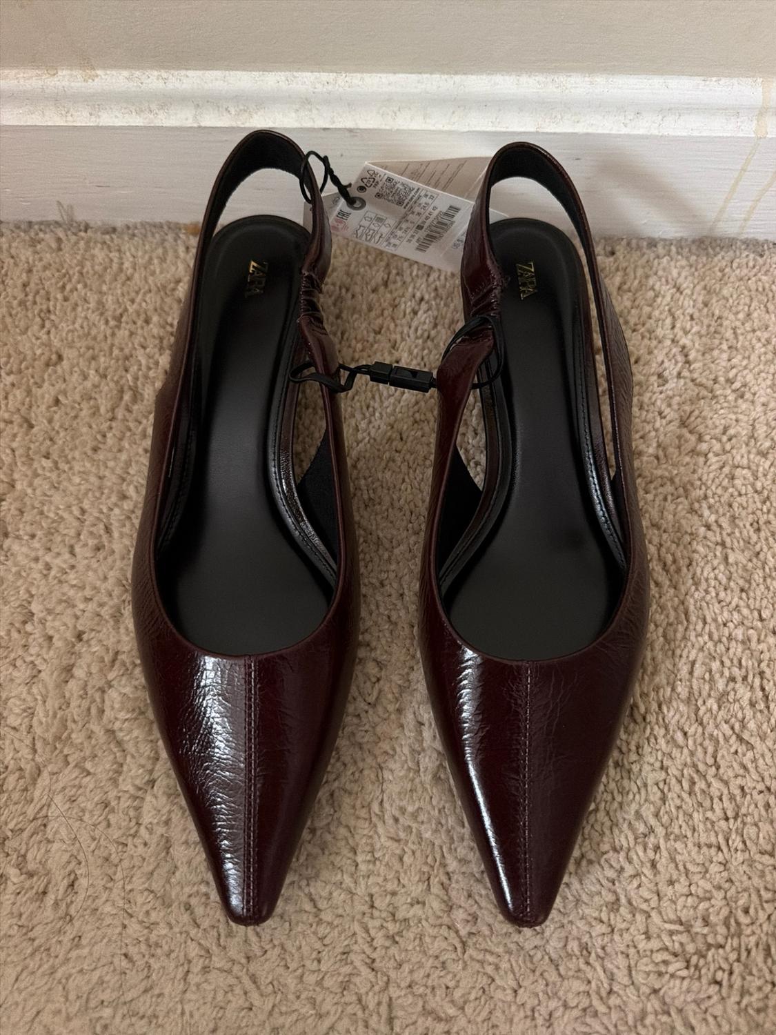 Pointed-toe backless shoes in shiny burgundy with the text ZARA on the insole, on a beige carpet.