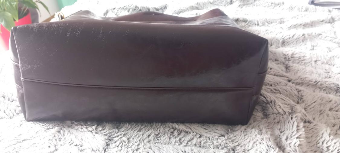 Base of a shiny dark brown handbag resting on a soft grey blanket. In the background, a green plant in a red pot can be seen.