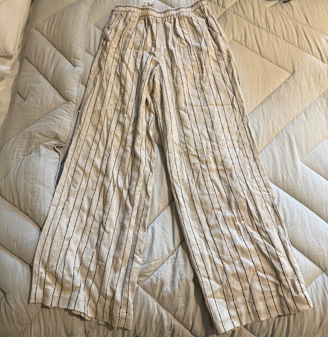 Wide-leg beige trousers with dark vertical stripes and an elasticated waist, laid out on a bed with a light-coloured duvet in a relaxed setting.