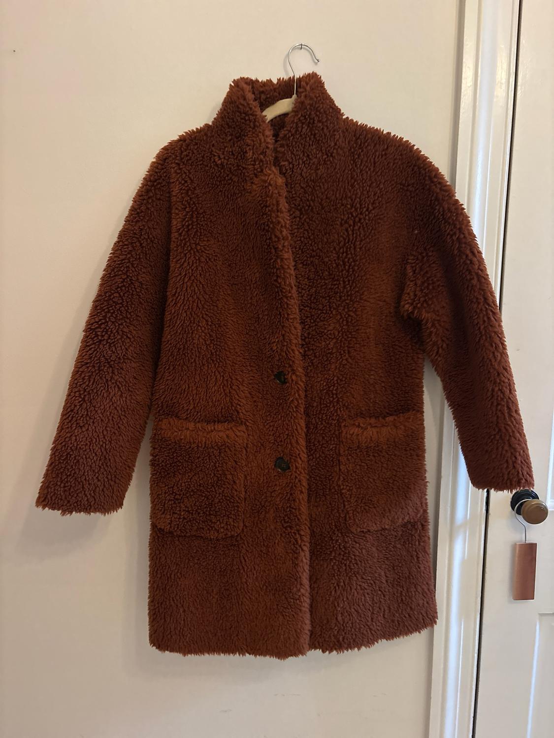 Curly-textured brown coat hanging on a hanger against a white wall.
Design with a high collar, two buttons, and front pockets.