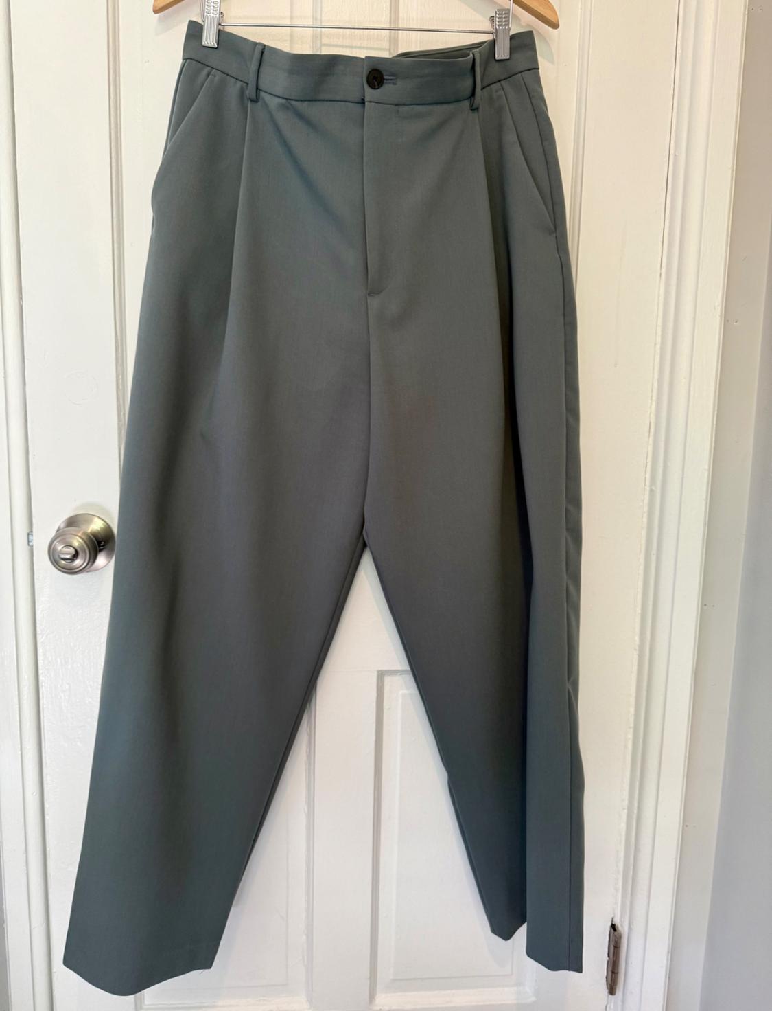 Gray pants hanging on a hanger over a white door. Classic design with pleats and a black button closure.
