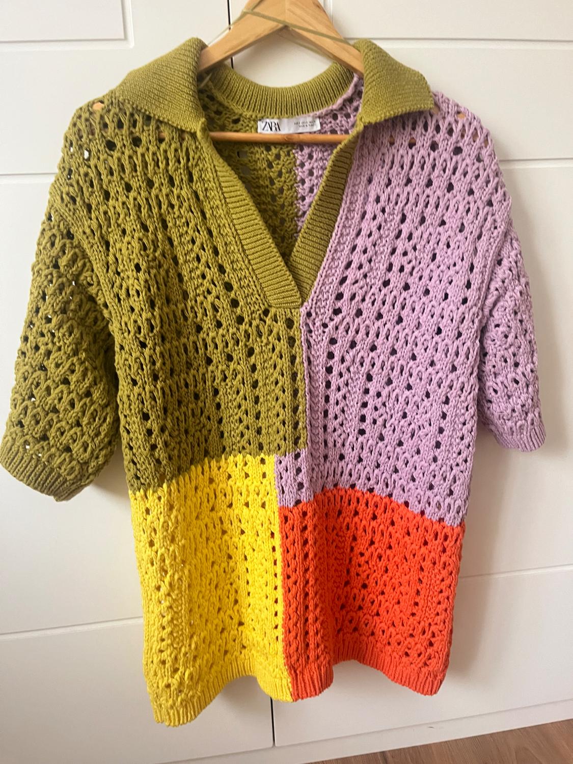 Crochet jumper in shades of green, lilac, yellow and orange, hanging on a wooden hanger against a white background.