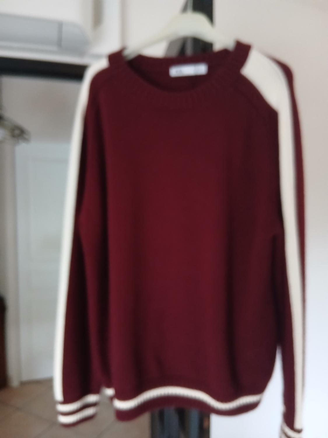 Burgundy jumper with white stripes on the sleeves and hem, hanging on a hanger in a blurred interior.
