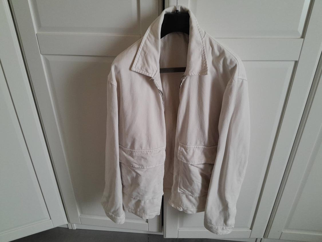 White jacket hanging on a hanger in front of a white wardrobe. The jacket has large pockets and a classic collar.