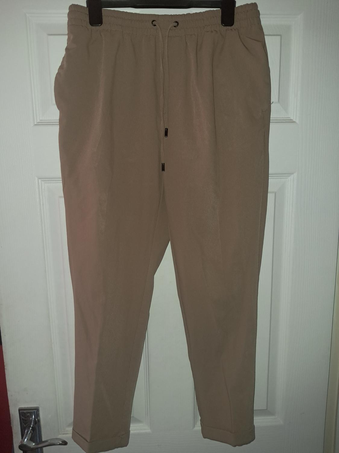Beige trousers with an elasticated drawstring waist, hanging on a black hanger in front of a white door. The side pockets stand out.